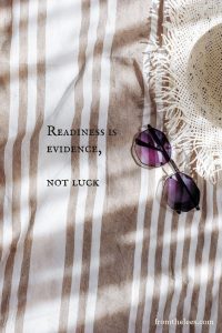 Purple-lensed sunglasses resting on a beige and white striped linen blanket beside the words, "Readiness is evidence, not luck." Warm sunlight and soft shadows create a calm, reflective scene that evokes preparation, confidence, and quiet clarity.