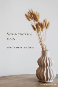 Decorative image featuring the quote "Satisfaction is a loop, not a destination" with a sculptural ceramic vase holding dried bunny tail grass on a wooden tabletop.